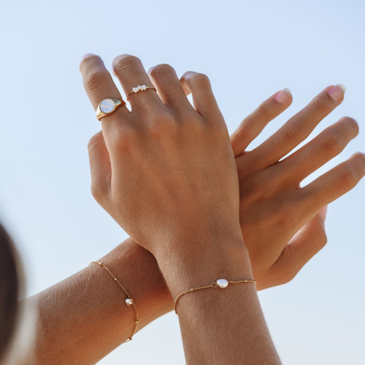 Dainty pearl rings and gold pearl bracelets worn on two hands against the sky